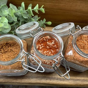 May include: Four glass jars with metal lids containing different types of spices. The jars are arranged in a row on a wooden surface. The spices are a light brown, a reddish-orange, and a darker brown.