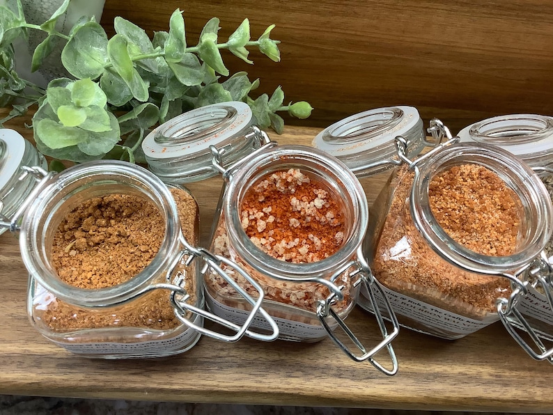May include: Four glass jars with metal lids containing different types of spices. The jars are arranged in a row on a wooden surface. The spices are a light brown, a reddish-orange, and a darker brown.