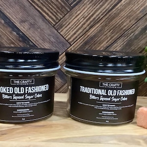 Puede incluir: Dos frascos de vidrio con tapas negras y etiquetas que dicen "SMOKED OLD FASHIONED" y "TRADITIONAL OLD FASHIONED" con "Bitters Infused Sugar Cubes". Al lado de los frascos hay cubos de azúcar. El fondo es una superficie de madera.
