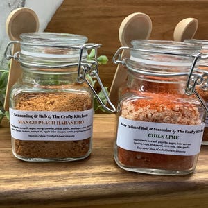 May include: Three glass jars with metal lids and wooden spoons. The jars contain different spice blends: Mango Peach Habanero, Chile Lime, and  Pecan Walnut. Each jar has a label with the name of the spice blend and the company name, The Crafty Kitchen.