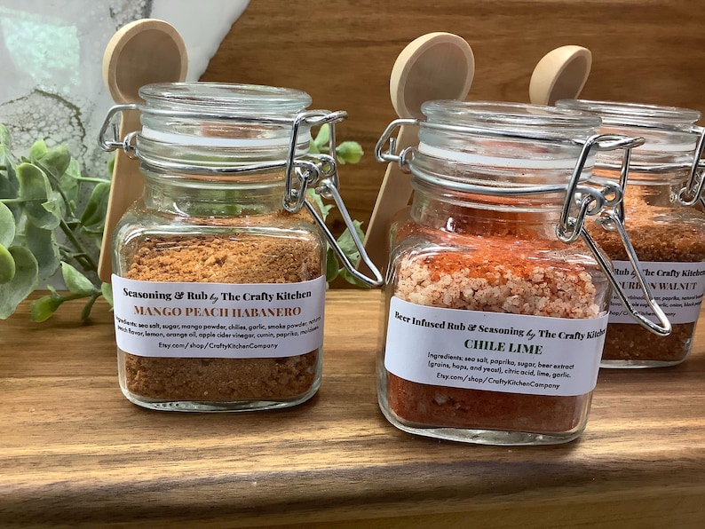 May include: Three glass jars with metal lids and wooden spoons. The jars contain different spice blends: Mango Peach Habanero, Chile Lime, and  Pecan Walnut. Each jar has a label with the name of the spice blend and the company name, The Crafty Kitchen.