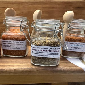 May include: Three glass jars with metal lids and labels containing different spice blends. The labels read "Maple Bourbon Walnut", "Maple Mesquite Peppercorn", and "Texas Two Step". The jars are on a wooden shelf.