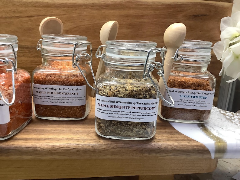 May include: Three glass jars with metal lids and labels containing different spice blends. The labels read "Maple Bourbon Walnut", "Maple Mesquite Peppercorn", and "Texas Two Step". The jars are on a wooden shelf.