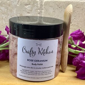 Op de afbeelding: Een glazen pot met een zwarte deksel gevuld met roze body scrub. De pot heeft een wit etiket met de tekst "THE Crafty Kitchen ROSE GERANIUM Body Polish" en gebruiksaanwijzingen. De pot staat op een houten oppervlak met paarse bloemen en een houten lepel.