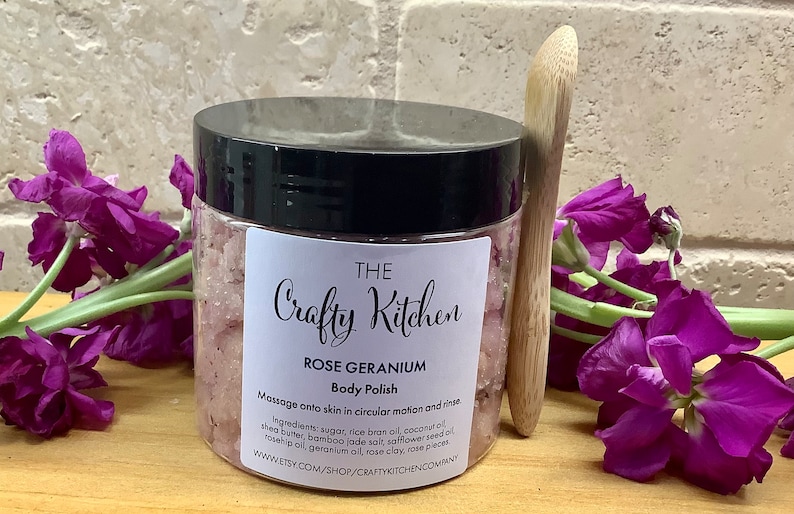 Op de afbeelding: Een glazen pot met een zwarte deksel gevuld met roze body scrub. De pot heeft een wit etiket met de tekst "THE Crafty Kitchen ROSE GERANIUM Body Polish" en gebruiksaanwijzingen. De pot staat op een houten oppervlak met paarse bloemen en een houten lepel.