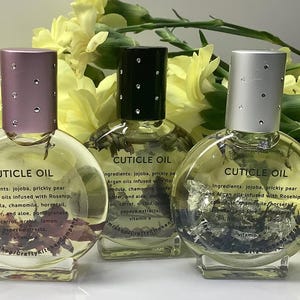 Op de afbeelding: Drie heldere, ronde nagelriemolieflessen met decoratieve deksels. De flessen bevatten zichtbare botanische elementen en de tekst "CUTICLE OIL" en informatie over de ingredi&euml;nten. De achtergrond heeft gele bloemen.