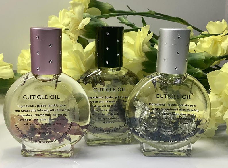 Op de afbeelding: Drie heldere, ronde nagelriemolieflessen met decoratieve deksels. De flessen bevatten zichtbare botanische elementen en de tekst "CUTICLE OIL" en informatie over de ingredi&euml;nten. De achtergrond heeft gele bloemen.