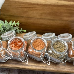 May include: Six glass jars with metal lids filled with different spices. The jars are arranged in a row on a wooden surface. The spices include red pepper flakes, salt, a blend of spices, and a dark spice.