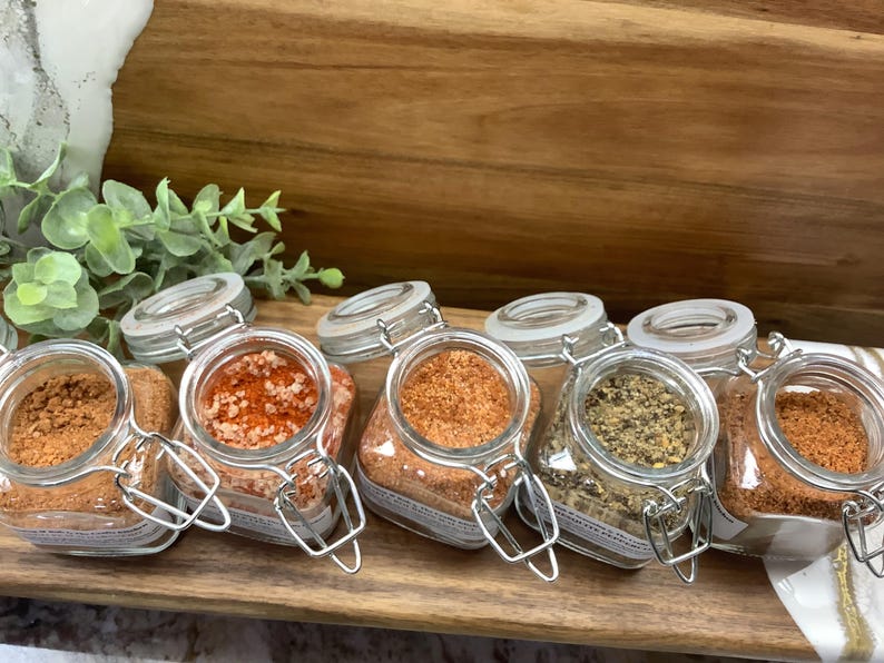 May include: Six glass jars with metal lids filled with different spices. The jars are arranged in a row on a wooden surface. The spices include red pepper flakes, salt, a blend of spices, and a dark spice.