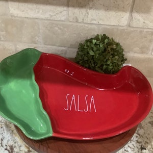 May include: Red and green ceramic salsa dish shaped like a chili pepper. The dish has a separate compartment for chips and the word "SALSA" is written on the red side.