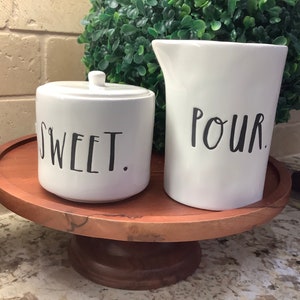 Puede incluir: Azucarero y jarra de crema de cerámica blanca con texto negro sobre un soporte de pastel de madera. El azucarero dice "SWEET." y la jarra de crema dice "POUR."