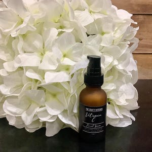 Puede incluir: Una botella de vidrio marr&oacute;n de suero facial de vitamina C con un dispensador de bomba negro. La etiqueta dice "The Crafty Kitchen" e incluye el nombre del producto y los ingredientes. La botella est&aacute; frente a un gran ramo de flores blancas.