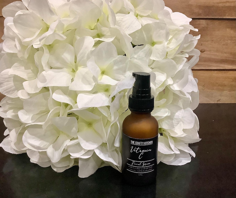 Puede incluir: Una botella de vidrio marr&oacute;n de suero facial de vitamina C con un dispensador de bomba negro. La etiqueta dice "The Crafty Kitchen" e incluye el nombre del producto y los ingredientes. La botella est&aacute; frente a un gran ramo de flores blancas.