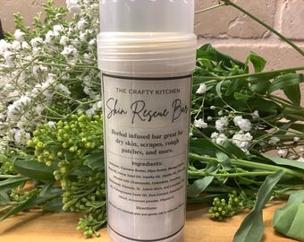 May include: A white plastic container with a label that reads "The Crafty Kitchen Skin Rescue Bar". The label describes the product as a herbal infused bar great for dry skin, scrapes, rough patches, and more. The ingredients are listed on the label.