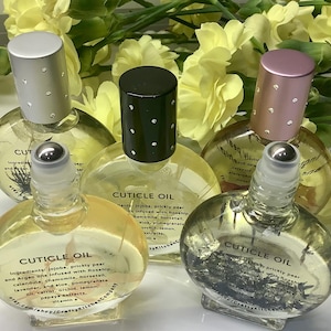 May include: Five clear glass cuticle oil bottles with silver, black, and pink caps. The bottles are labeled with the words "CUTICLE OIL" and ingredient information. Yellow flowers are in the background.