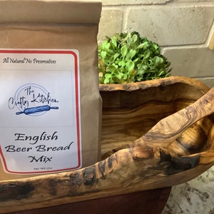 Pode incluir: Um saco de papel castanho com um rótulo que diz "The Crafty Kitchen" e "English Beer Bread Mix". O saco está numa tigela de madeira com uma colher de madeira.