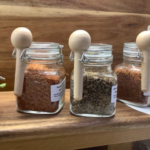 May include: Four glass jars with wooden spoons attached to the lids, filled with different spices. The jars are labeled "Chili Lime", "Seasoning", "Montreal", and "Rub & Seasoning".