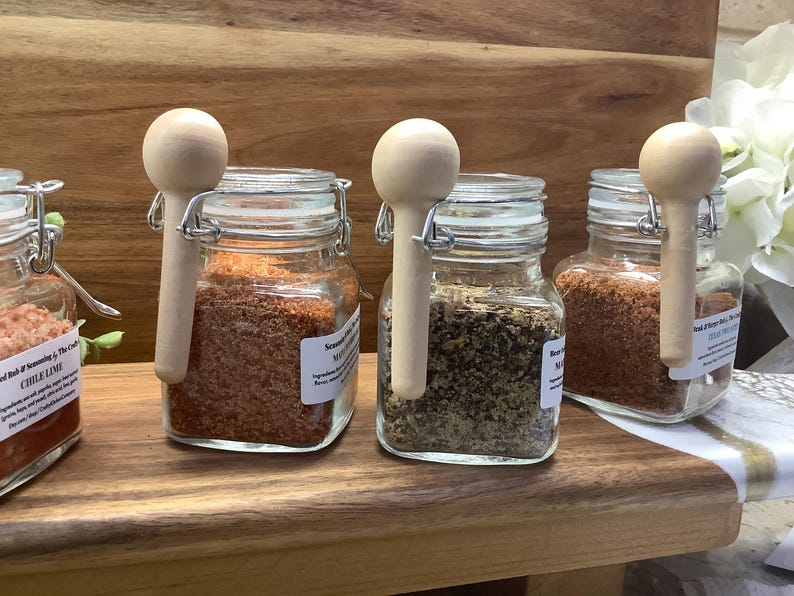 May include: Four glass jars with wooden spoons attached to the lids, filled with different spices. The jars are labeled "Chili Lime", "Seasoning", "Montreal", and "Rub & Seasoning".