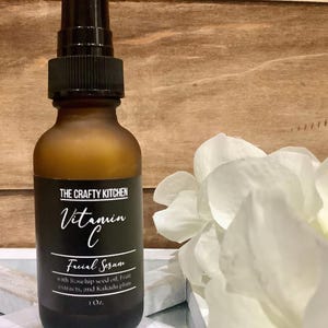 Puede incluir: Una botella de vidrio marr&oacute;n de suero facial de vitamina C de The Crafty Kitchen. La etiqueta es negra con texto blanco, incluyendo "Vitamin C" y "Facial Serum". La botella contiene 29.57 ml de suero y est&aacute; junto a flores blancas.