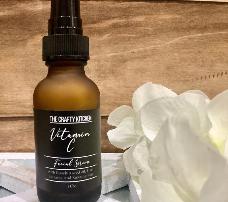 Puede incluir: Una botella de vidrio marr&oacute;n de suero facial de vitamina C de The Crafty Kitchen. La etiqueta es negra con texto blanco, incluyendo "Vitamin C" y "Facial Serum". La botella contiene 29.57 ml de suero y est&aacute; junto a flores blancas.