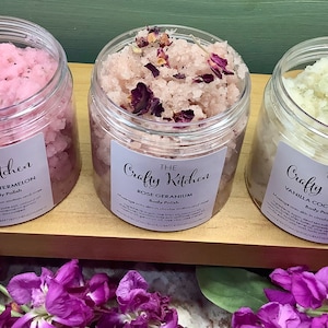 Op de afbeelding: Drie glazen potten met lichaamsscrub met etiketten waarop "The Crafty Kitchen" staat. De potten zijn gevuld met roze, witte en roze scrubs. De etiketten geven de geuren aan als "Aardbei-Watermeloen", "Roos-Geranium" en "Vanille-Cacaoboter".