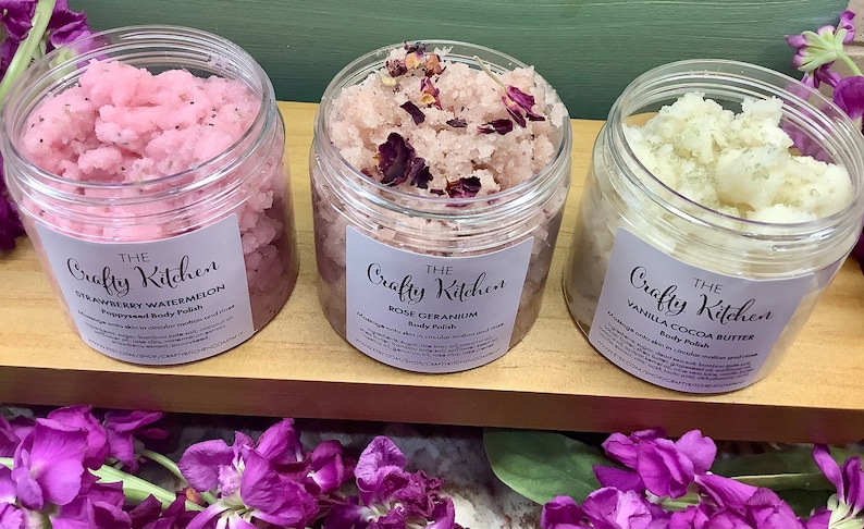 Op de afbeelding: Drie glazen potten met lichaamsscrub met etiketten waarop "The Crafty Kitchen" staat. De potten zijn gevuld met roze, witte en roze scrubs. De etiketten geven de geuren aan als "Aardbei-Watermeloen", "Roos-Geranium" en "Vanille-Cacaoboter".