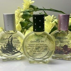 Op de afbeelding: Drie heldere glazen nagelriemolieflessen met zilveren, zwarte en roze doppen. Elke fles heeft een ander ontwerp en het opschrift "CUTICLE OIL". Gele bloemen op de achtergrond.