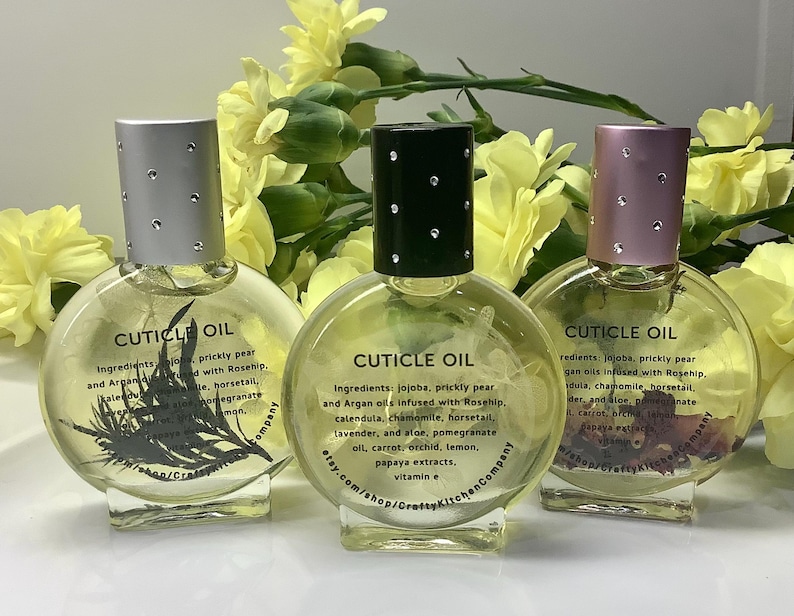 Op de afbeelding: Drie heldere glazen nagelriemolieflessen met zilveren, zwarte en roze doppen. Elke fles heeft een ander ontwerp en het opschrift "CUTICLE OIL". Gele bloemen op de achtergrond.