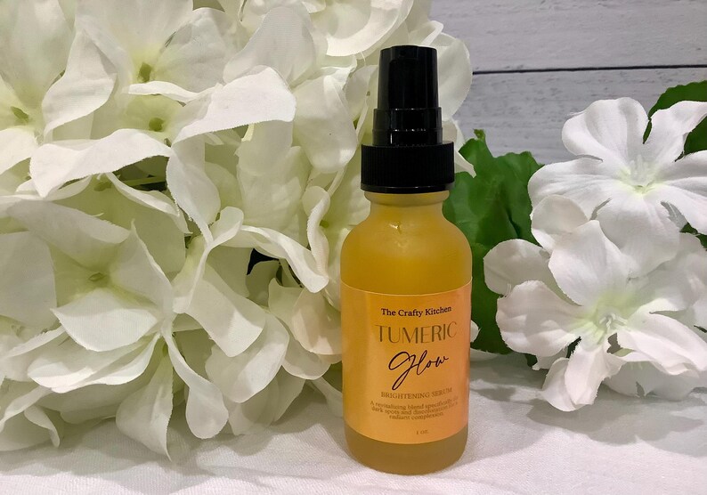 Puede incluir: Una botella de vidrio amarillo con una tapa de bomba negra, etiquetada como "Tumeric Glow Brightening Serum", se encuentra frente a flores blancas. La botella est&aacute; rodeada de flores blancas y hojas verdes.