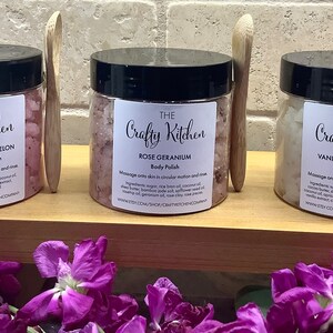 Op de afbeelding: Drie glazen potten met lichaamsscrub en houten lepels. De potten zijn gelabeld met "The Crafty Kitchen" en hebben verschillende geuren: Aardbei-Watermeloen-Lichaams Scrub met Papaverzaad, Rozen-Geranium-Lichaams Scrub en Vanille-Cacao Boter Lichaams Scrub.