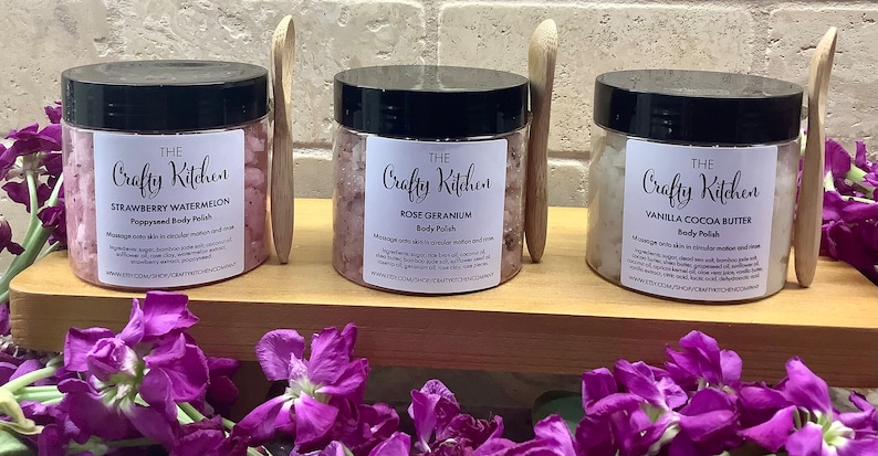Op de afbeelding: Drie glazen potten met lichaamsscrub en houten lepels. De potten zijn gelabeld met "The Crafty Kitchen" en hebben verschillende geuren: Aardbei-Watermeloen-Lichaams Scrub met Papaverzaad, Rozen-Geranium-Lichaams Scrub en Vanille-Cacao Boter Lichaams Scrub.