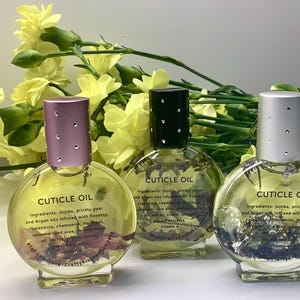 Op de afbeelding: Drie heldere glazen flessen nagelriemolie met decoratieve deksels. Elke fles bevat een heldere vloeistof met zichtbare botanische elementen. De flessen zijn voorzien van het label "CUTICLE OIL" en worden omringd door gele bloemen.