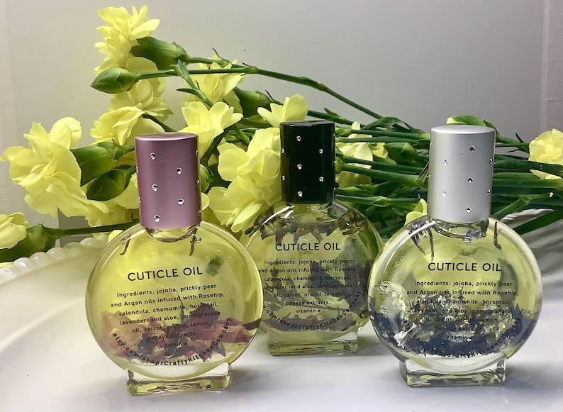 Op de afbeelding: Drie heldere glazen flessen nagelriemolie met decoratieve deksels. Elke fles bevat een heldere vloeistof met zichtbare botanische elementen. De flessen zijn voorzien van het label "CUTICLE OIL" en worden omringd door gele bloemen.