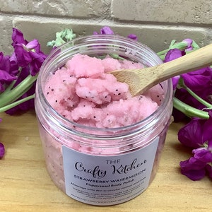 Op de afbeelding: Een transparante pot met roze aardbei-watermeloen body scrub met een houten lepel die het product opschept. De pot heeft een wit etiket met de tekst "The Crafty Kitchen Strawberry Watermelon Papaya Body Polish".