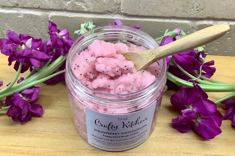 Op de afbeelding: Een transparante pot met roze aardbei-watermeloen body scrub met een houten lepel die het product opschept. De pot heeft een wit etiket met de tekst "The Crafty Kitchen Strawberry Watermelon Papaya Body Polish".