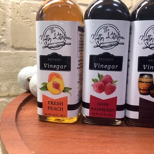 May include: Three glass bottles of balsamic vinegar with labels featuring the text "The Crafty Kitchen". The bottles are labeled with different flavors: "Fresh Peach", "Dark Raspberry", and "Traditional". Each bottle contains 60 ml (2 fl oz) of vinegar.