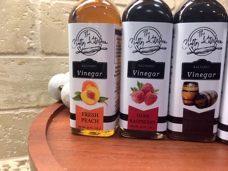 May include: Three glass bottles of balsamic vinegar with labels featuring the text "The Crafty Kitchen". The bottles are labeled with different flavors: "Fresh Peach", "Dark Raspberry", and "Traditional". Each bottle contains 60 ml (2 fl oz) of vinegar.