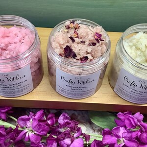 Op de afbeelding: Drie glazen potten met lichaamsscrub met etiketten waarop "The Crafty Kitchen" staat. De potten zijn gevuld met roze, rozekleurige en witte scrubs. De potten staan op een houten oppervlak.