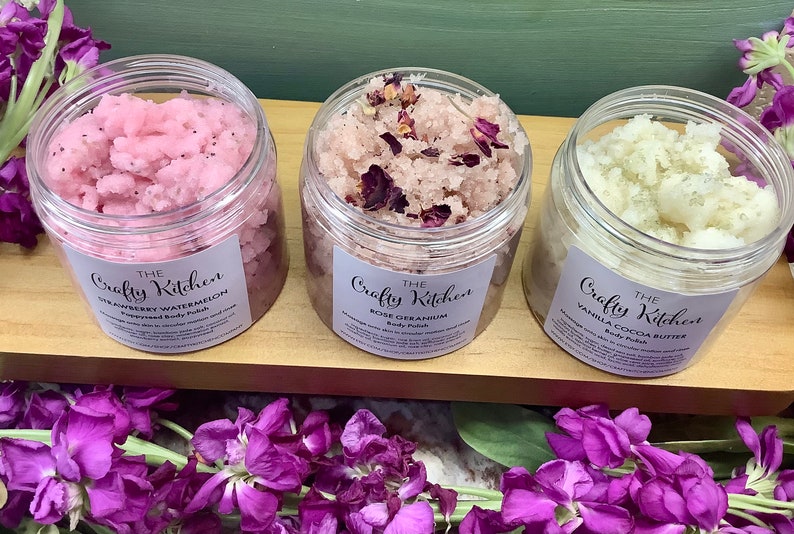 Op de afbeelding: Drie glazen potten met lichaamsscrub met etiketten waarop "The Crafty Kitchen" staat. De potten zijn gevuld met roze, rozekleurige en witte scrubs. De potten staan op een houten oppervlak.