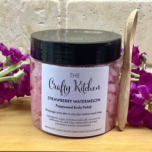 Op de afbeelding: Een glazen pot met roze aardbei-watermeloen-papaverzaad-lichaamspoeder met een houten lepel. De pot heeft een zwarte deksel en een wit etiket met de tekst "THE Crafty Kitchen STRAWBERRY WATERMELON Poppyseed Body Polish Massage onto skin in circular motion and rinse. Ingredients: sugar, bamboo jade salt, coconut oil, safflower oil, rose clay, watermelon extract, strawberry extract, poppyseed. www.ETSY.COM/SHOP/CRAFTYKITCHENCOMPANY"