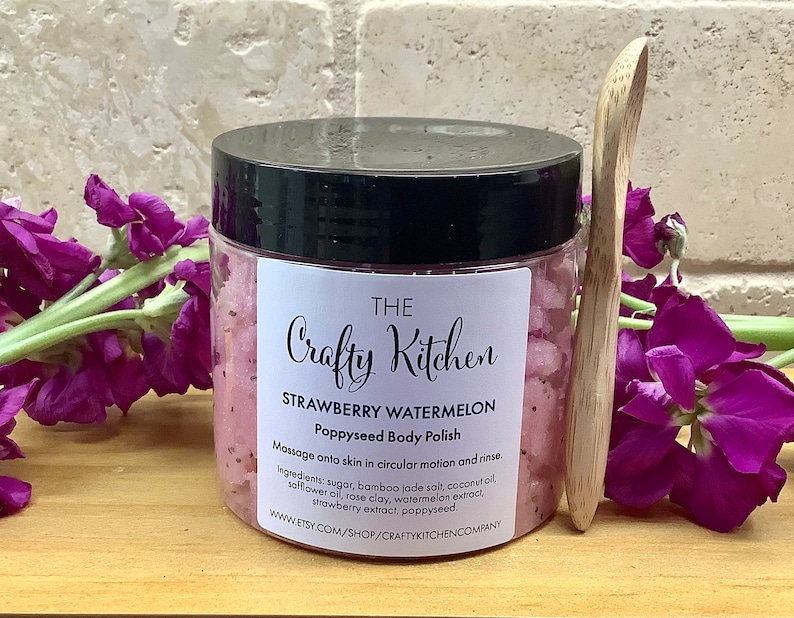 Op de afbeelding: Een glazen pot met roze aardbei-watermeloen-papaverzaad-lichaamspoeder met een houten lepel. De pot heeft een zwarte deksel en een wit etiket met de tekst "THE Crafty Kitchen STRAWBERRY WATERMELON Poppyseed Body Polish Massage onto skin in circular motion and rinse. Ingredients: sugar, bamboo jade salt, coconut oil, safflower oil, rose clay, watermelon extract, strawberry extract, poppyseed. www.ETSY.COM/SHOP/CRAFTYKITCHENCOMPANY"