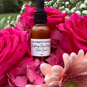 May include: A brown glass bottle with a dropper top, labeled "The Crafty Kitchen Raspberry Rose Hips Cream Serum 1 oz.", surrounded by pink and white flowers.