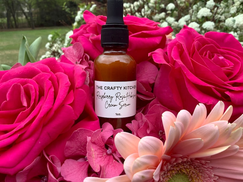 May include: A brown glass bottle with a dropper top, labeled "The Crafty Kitchen Raspberry Rose Hips Cream Serum 1 oz.", surrounded by pink and white flowers.