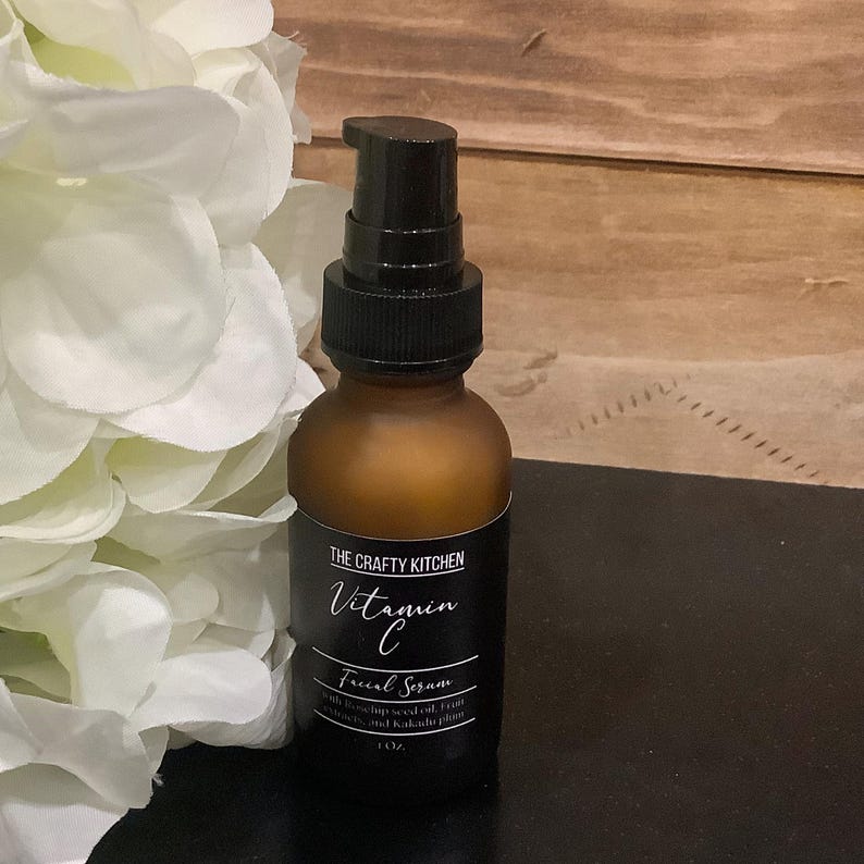 Puede incluir: Una botella de vidrio marr&oacute;n con un dispensador de bomba negro. La etiqueta dice "THE CRAFTY KITCHEN Vitamin C Facial Serum" y enumera los ingredientes. La botella es de 30 ml. Flores blancas en el fondo.