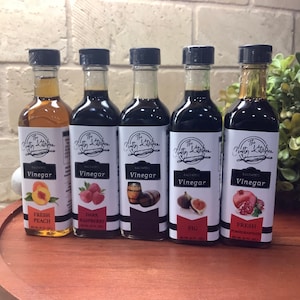 May include: Five glass bottles of balsamic vinegar with labels that say "The Nifty Kitchen" and the flavor of each vinegar. The flavors are Fresh Peach, Dark Raspberry, Oak Barrel, Fig, and Fresh Pomegranate.