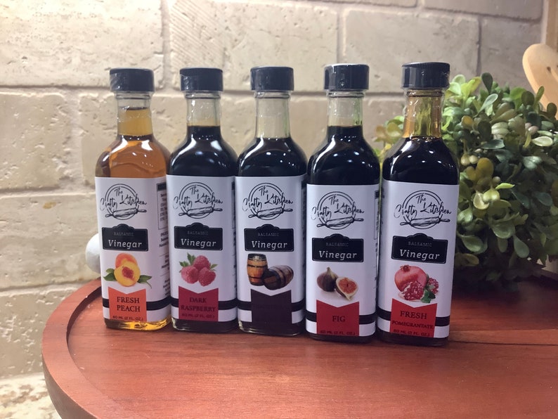 May include: Five glass bottles of balsamic vinegar with labels that say "The Nifty Kitchen" and the flavor of each vinegar. The flavors are Fresh Peach, Dark Raspberry, Oak Barrel, Fig, and Fresh Pomegranate.
