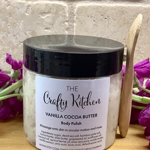 Op de afbeelding: Een witte pot met een zwarte deksel met het label "THE Crafty Kitchen VANILLA COCOA BUTTER Body Polish". De pot is gevuld met een witte, korrelige substantie. Het etiket bevat instructies om het product in cirkelvormige bewegingen op de huid te masseren en af te spoelen. De ingredi&euml;nten staan ​​onder de instructies vermeld. Een houten lepel bevindt zich naast de pot.