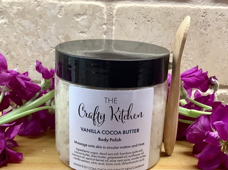 Op de afbeelding: Een witte pot met een zwarte deksel met het label "THE Crafty Kitchen VANILLA COCOA BUTTER Body Polish". De pot is gevuld met een witte, korrelige substantie. Het etiket bevat instructies om het product in cirkelvormige bewegingen op de huid te masseren en af te spoelen. De ingredi&euml;nten staan ​​onder de instructies vermeld. Een houten lepel bevindt zich naast de pot.
