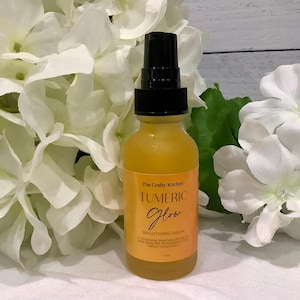 Puede incluir: Una botella de vidrio amarilla con un pulverizador negro, etiquetada como "Tumeric Glow Brightening Serum", se encuentra frente a flores blancas. La etiqueta de la botella es naranja con texto negro y dorado. El fondo incluye flores blancas y hojas verdes.