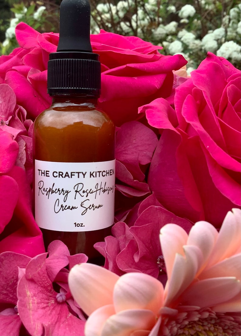 May include: A brown glass bottle with a dropper top, containing a clear liquid. The bottle is labeled "The Crafty Kitchen Raspberry Rose Hibiscus Cream Serum 1 oz.". The bottle is surrounded by pink and white flowers.